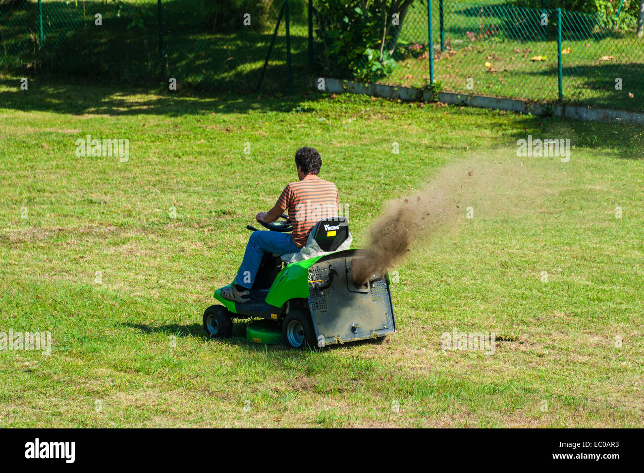 A man on a ride-on mower, cutting the grass without using a grass box and so throwing dust and stones into the air. Stock Photo