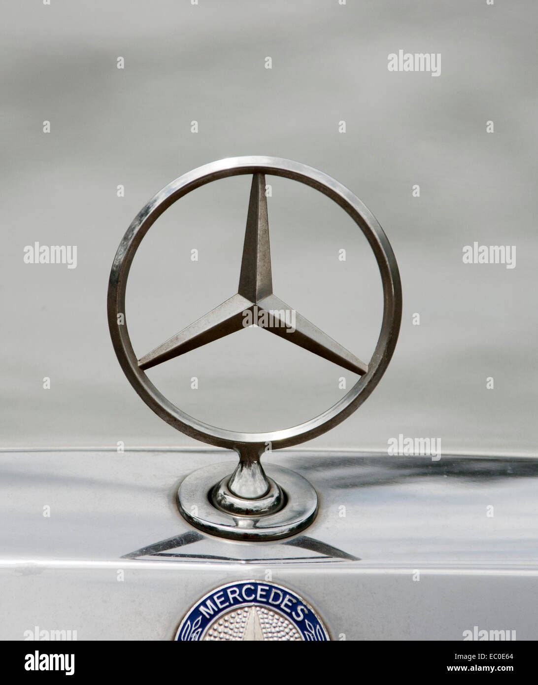 Mercedes silver metallic car bonnet emblem against  pale grey background, with manufacturer's name on shiny silver panel below Stock Photo