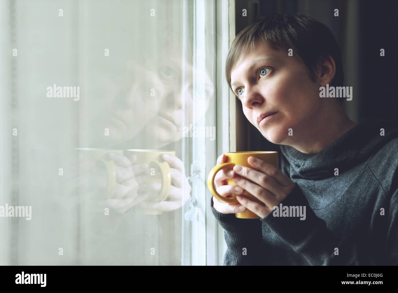 Lonely woman drinking cup of coffee by the window of her living room, looking out with a sad look on her face. Selective focus w Stock Photo