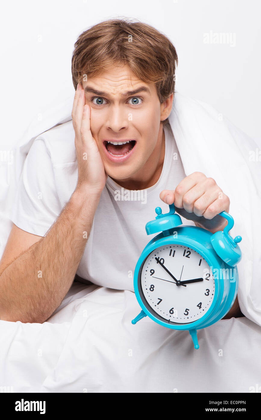 Young handsome man screaming and holding blue clock after oversleeping