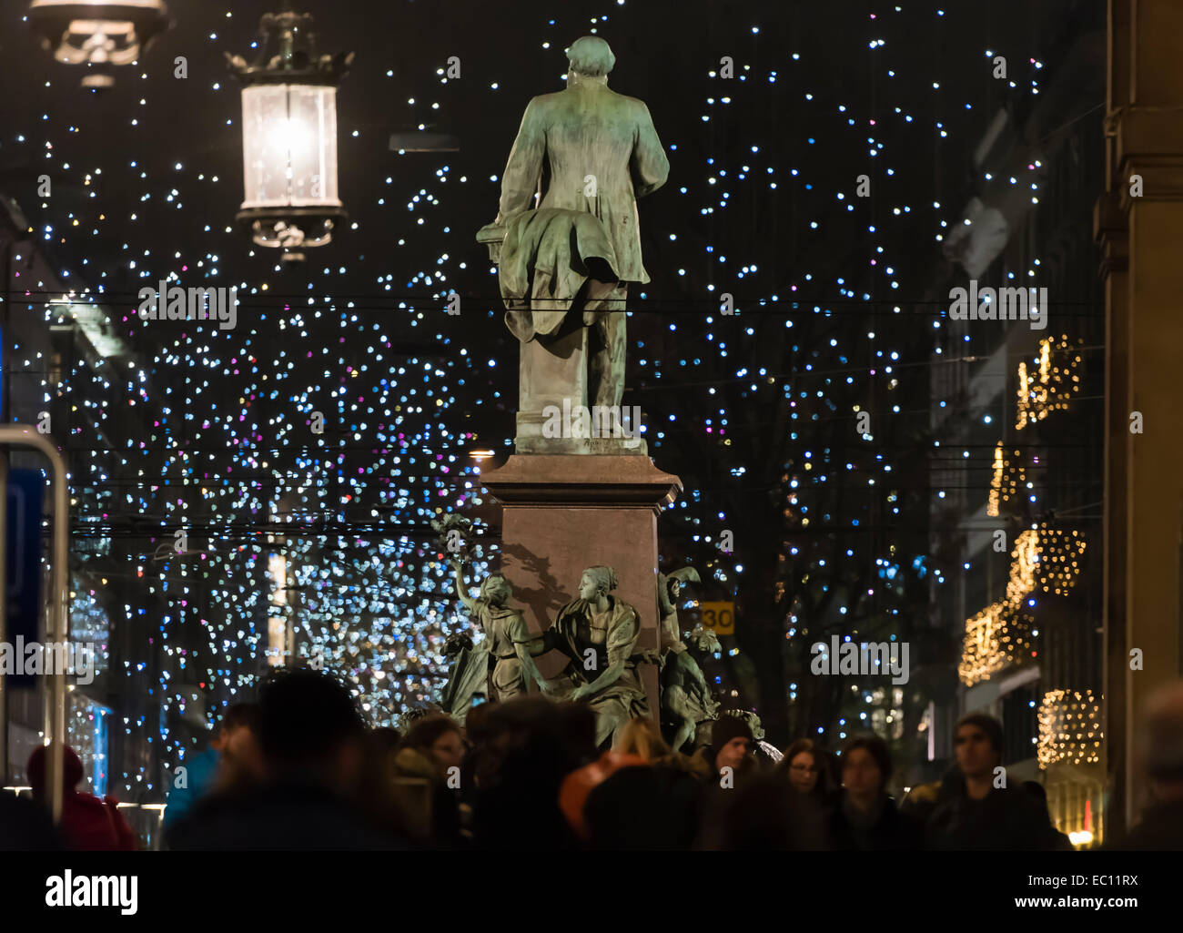 People at Zurich main station move past the statue of Alfred Escher with the city's Christmas illumination glowing in the back. Stock Photo