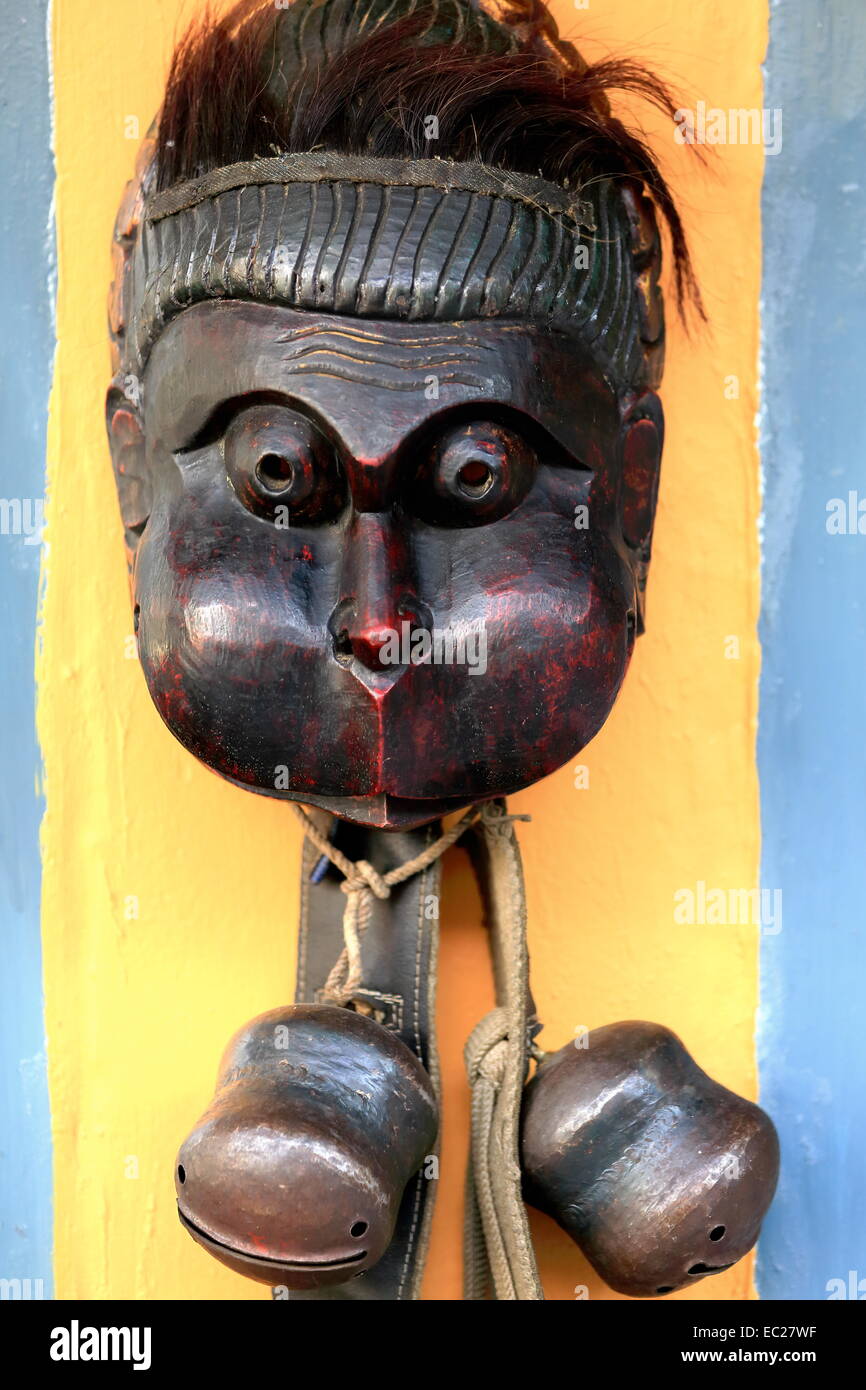 Traditional nepalese wooden mask with hair-swollen cheeks-rattles hanging from leather and fabric straps wall hanging on yellow Stock Photo