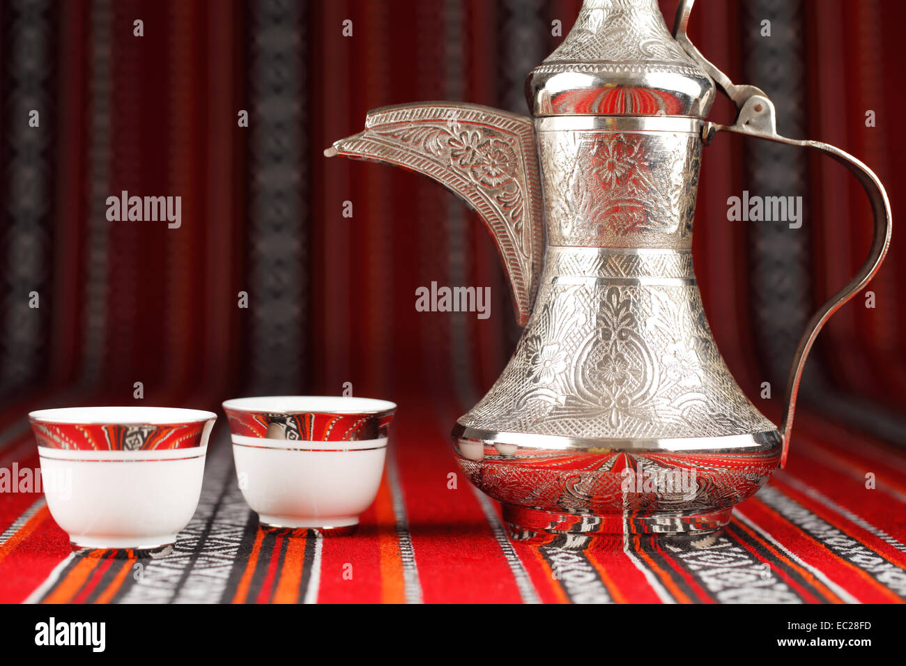 Ornate Arabian tea cups and a dallah are placed on traditional red fabric from the Gulf region. Stock Photo