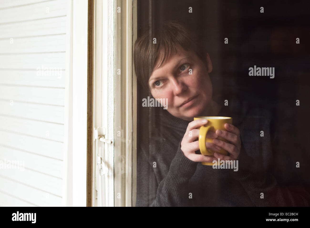 Lonely woman drinking cup of coffee by the window of her living room, looking out with a sad look on her face. Selective focus w Stock Photo
