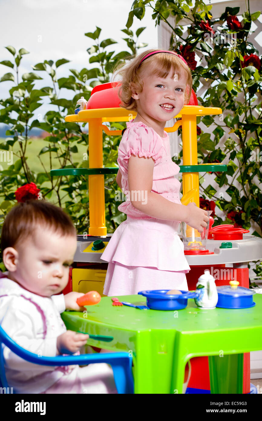 a 6 month old baby and a 4 years old girl in front of toy kitchen Stock Photo