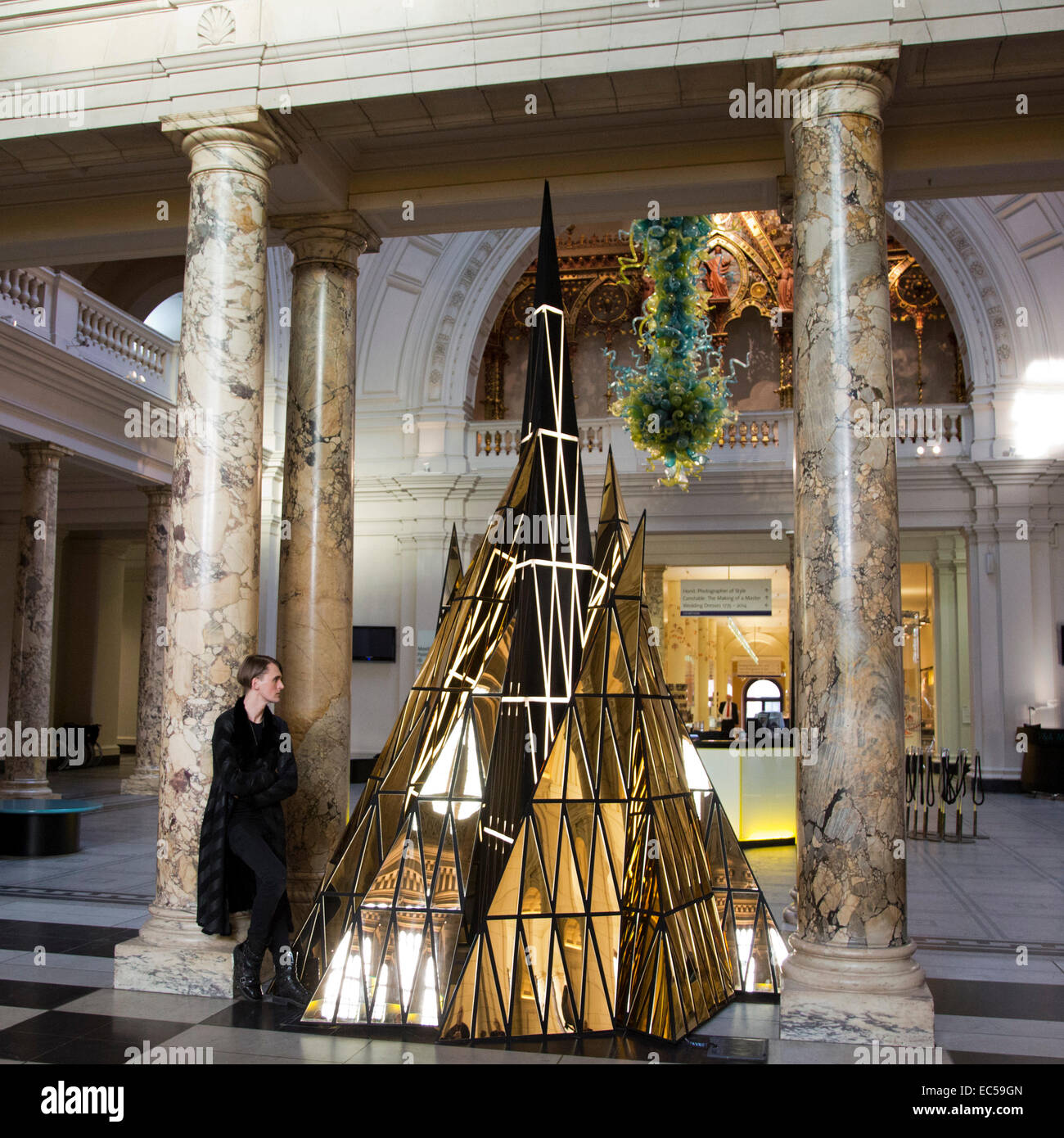 London, UK. 9 December 2014. British fashion designer Gareth Pugh has been commissioned to create a Christmas installation for the Grand Entrance of the V&A. 'Ceremony' stands at over 4m high and mirrors the shape of the traditional Christmas evergreen tree. Nine tiered pyramids are clad in gold which collect around a central beacon of light to represent an abstract nativity. Various materials have been used in the tree's creation including wood, fabric, acrylic and LED lights. Visitors to the V&A Museum can see the Christmas tree from 9 December 2014 to 6 January 2015. Stock Photo