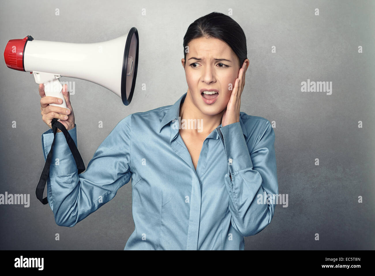 Young woman holding a megaphone to her head and clutching her ear with an aggrieved expression at the loudness of the sound so c Stock Photo