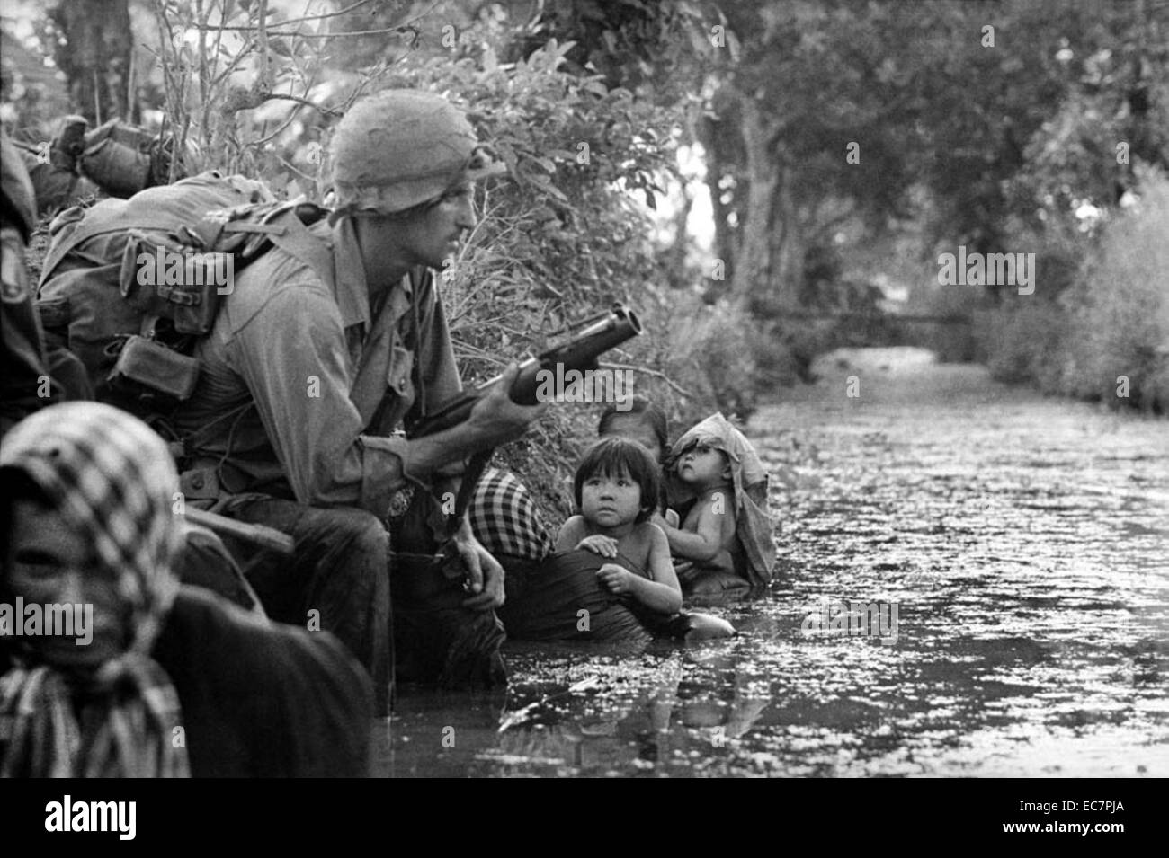 Photograph of two South Vietnamese children gazing at an American paratrooper holding an M79 grenade launcher as they cling to their mothers who huddle against a canal bank for protection from sniper fire in the Bao Trai area, Vietnam, 1966 Stock Photo