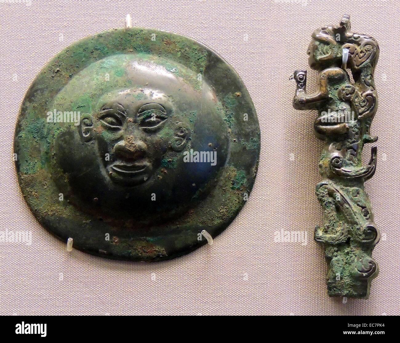 Left: Bronze circular fitting (From a shield or harness), depicting a human face.  Right: Bronze fitting depicting a tiger and a man who is holding an owl.. Western Zhou period, Chinese, 11-10th century BC Stock Photo