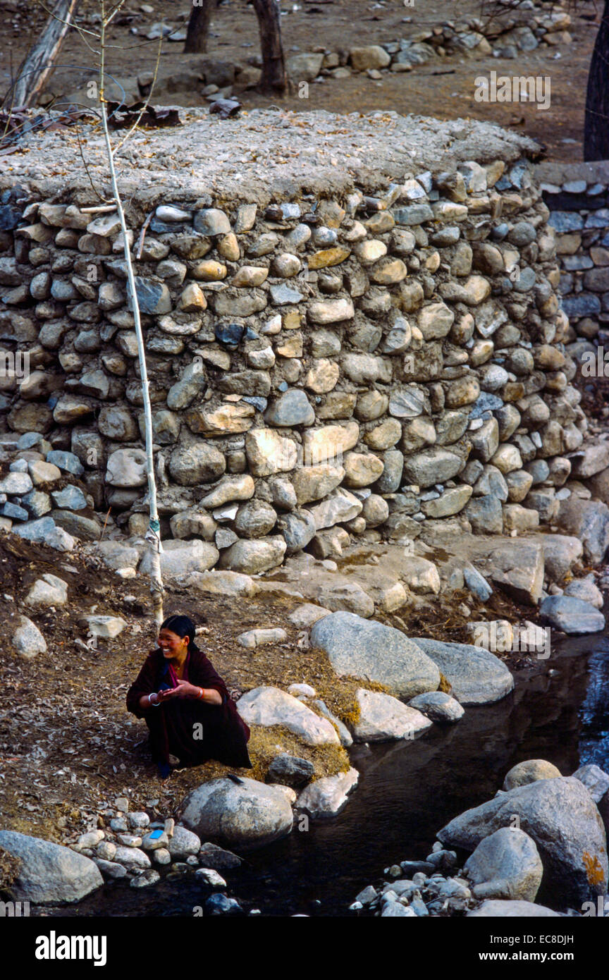 Ladakh Tibet woman smiling crouching washing clothes rubble wall stones black water Stock Photo