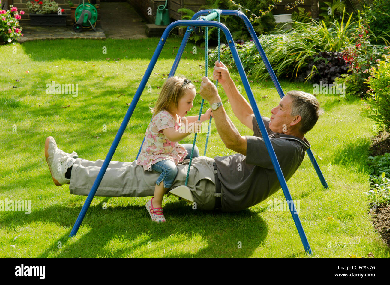Grandfather and granddaughter sitting on swing in garden. England, UK. September Stock Photo