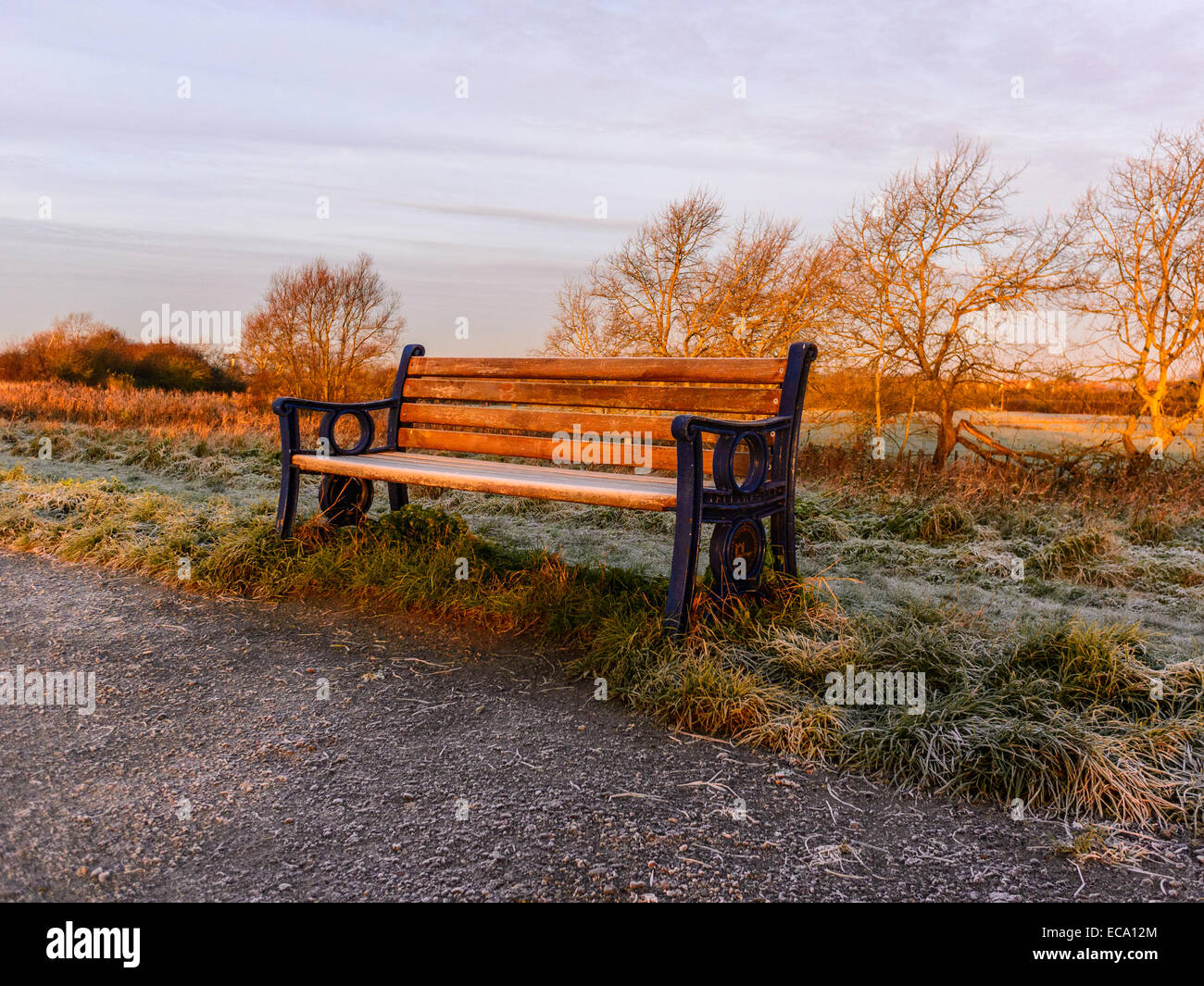 Countryside scene with early morning frost covering an empty frozen park bench with the surrounding area light up by the sunrise Stock Photo