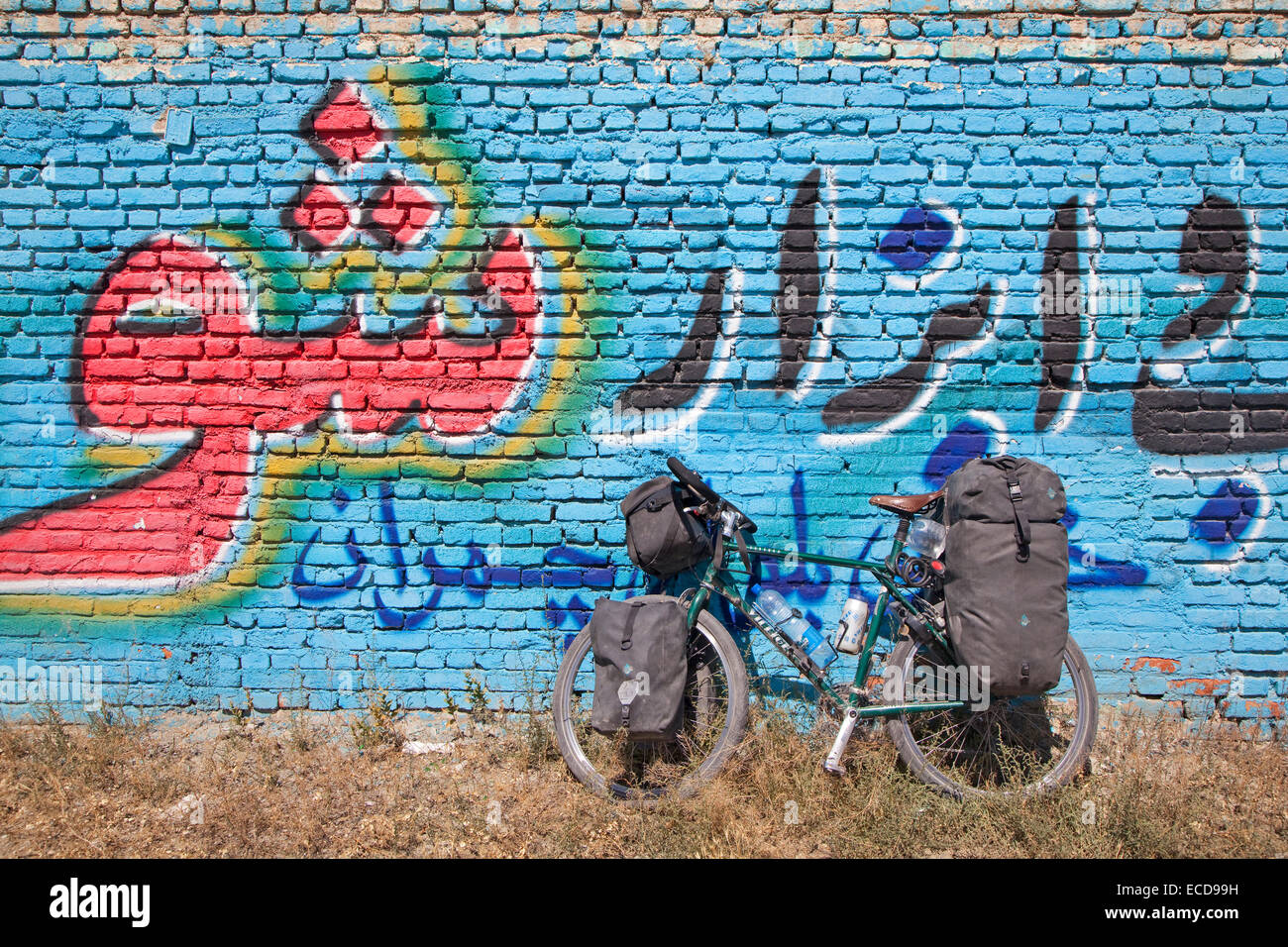 Touring bicycle equipped with water bottles in cages, four panniers and a handlebar bag in front of graffiti covered wall, Iran Stock Photo