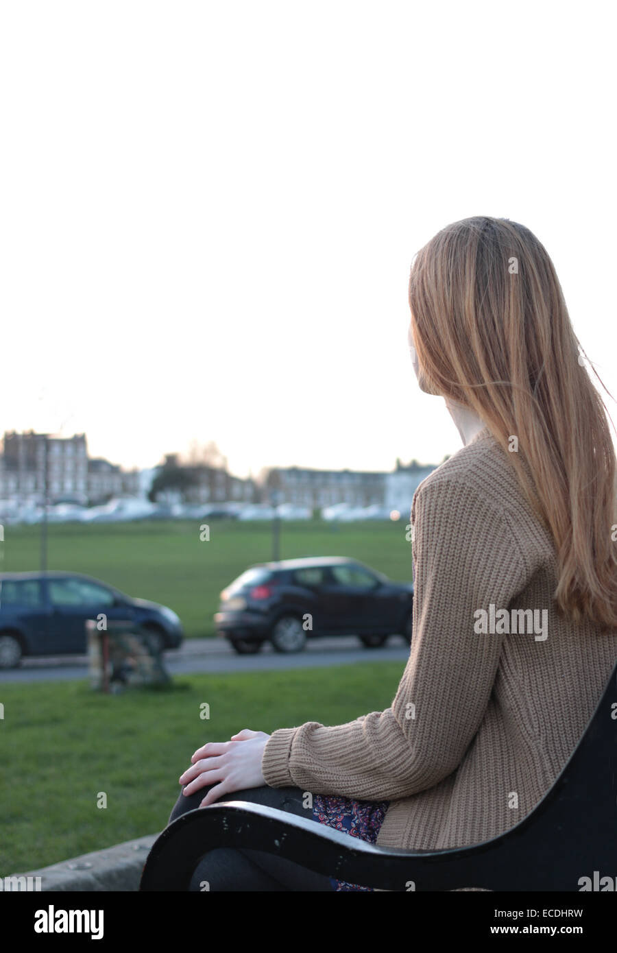 Rear view shot of a young woman sitting on a bench, looking into the distance in a park with cars parked. Stock Photo