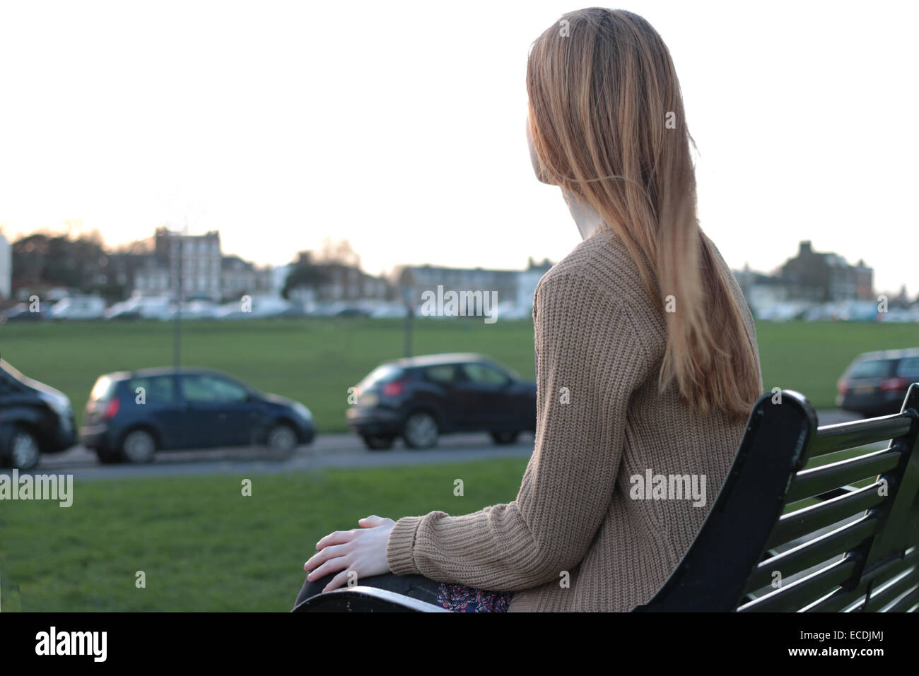 Rear view shot of a young woman sitting on a bench, looking into the distance in a park with cars parked. Stock Photo