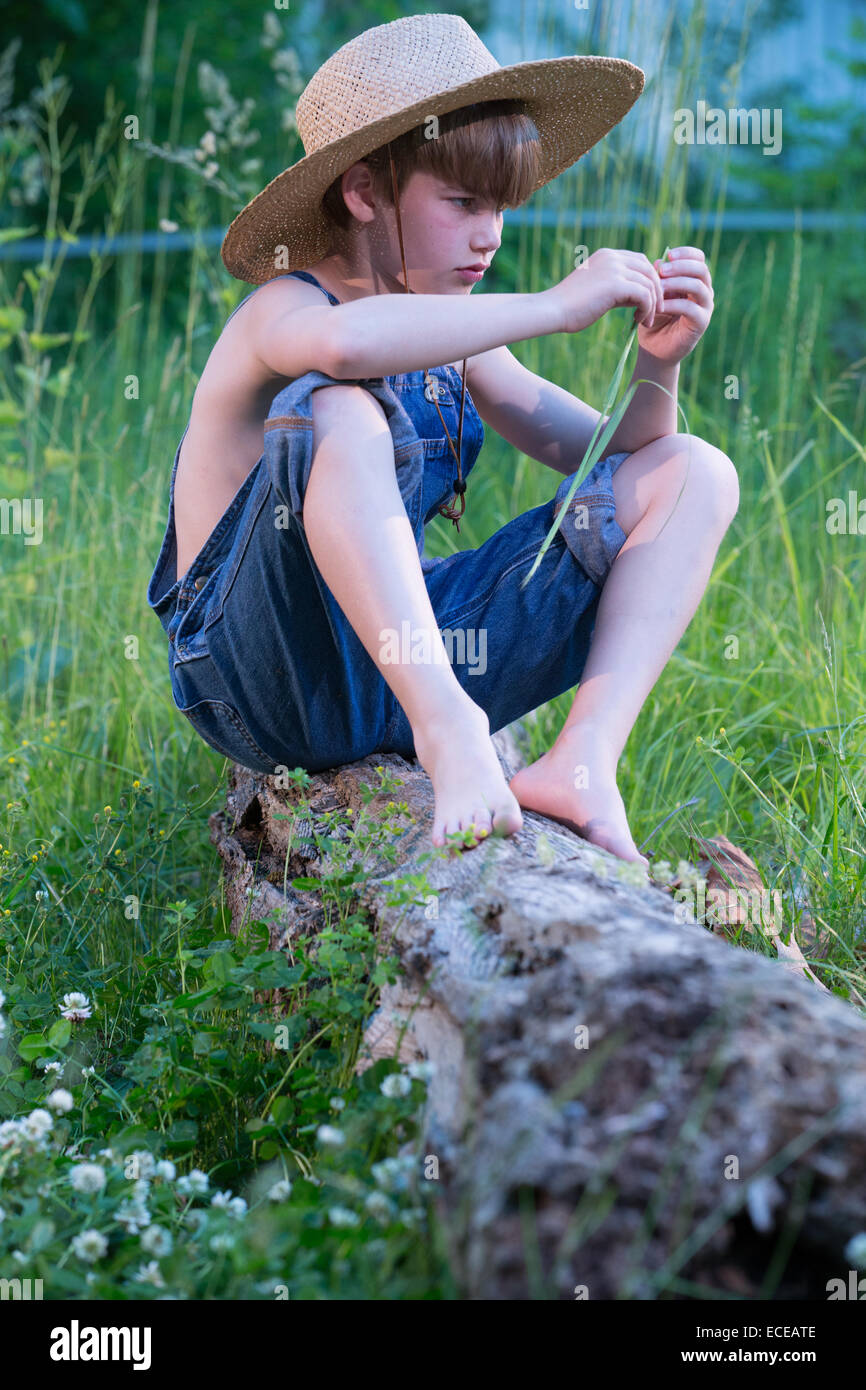 Young boy wearing blue overalls sitting on fallen tree Stock Photo