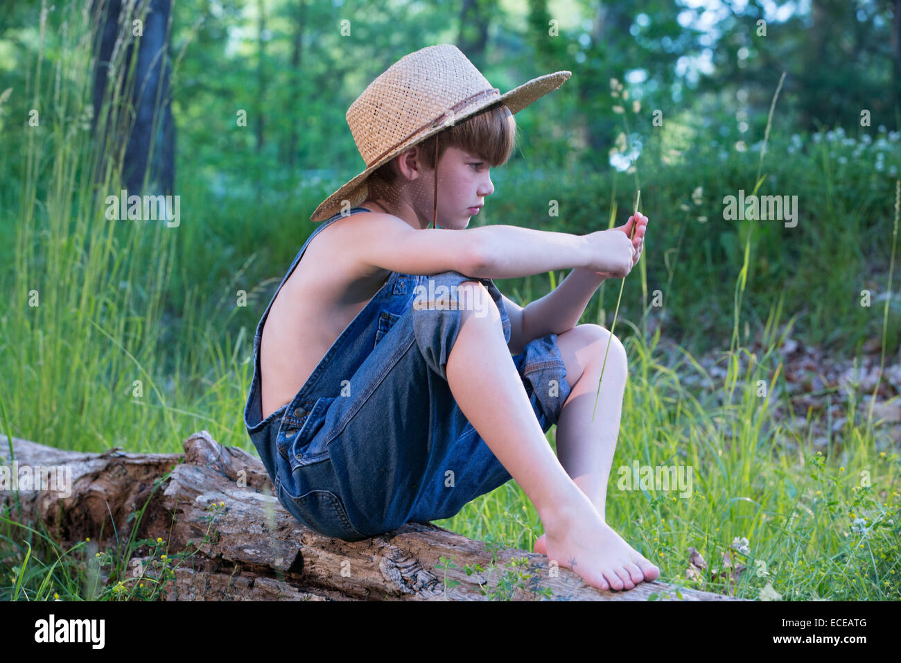 Young boy wearing straw hat sitting on log Stock Photo