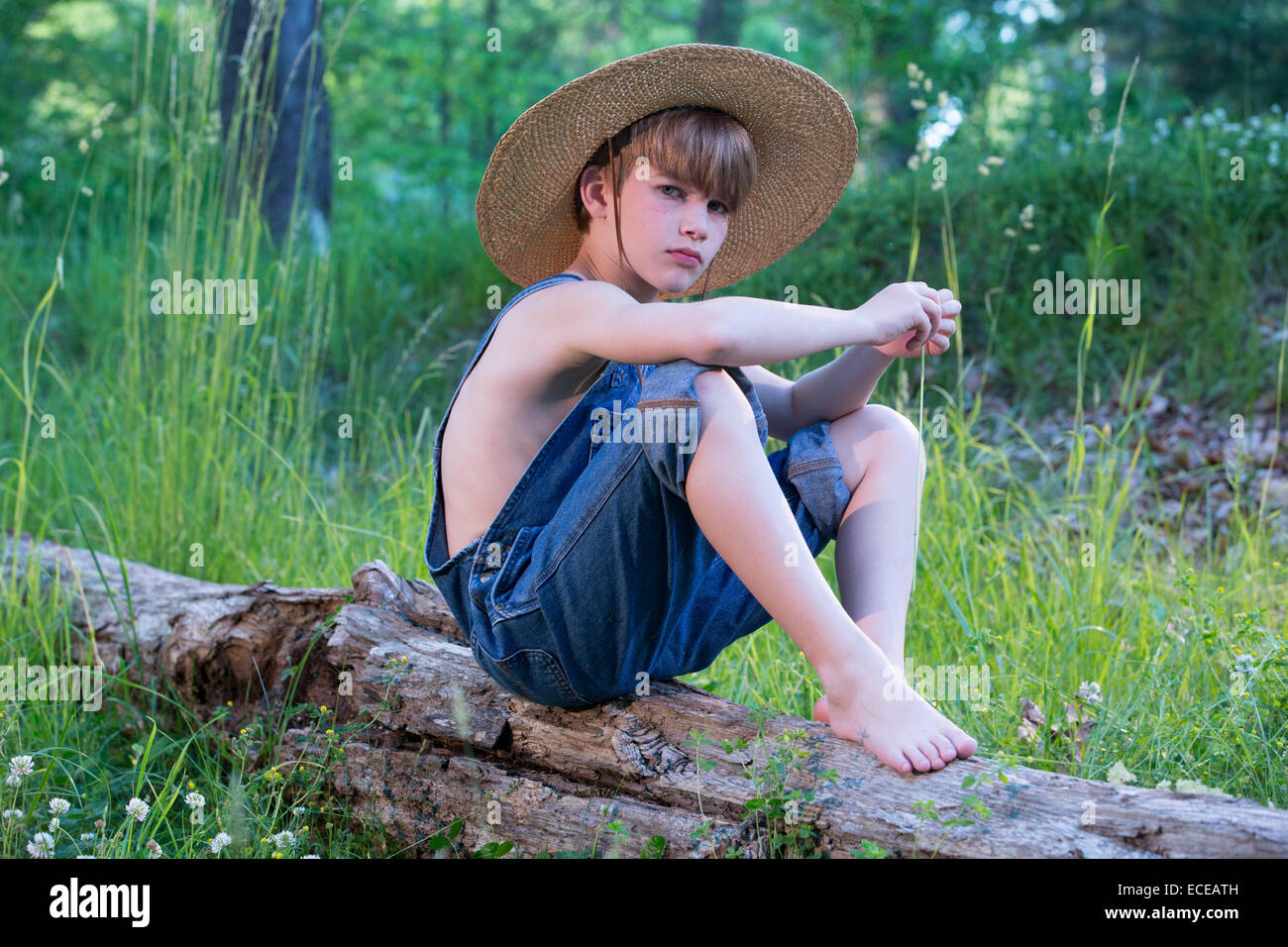 Young boy sitting on log wearing straw hat and blue overalls Stock Photo