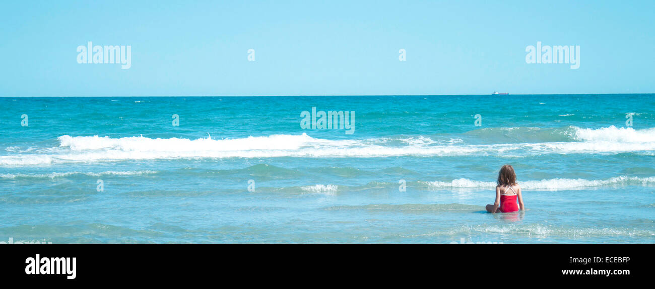 Rear view of a girl sitting on beach at water's edge Stock Photo