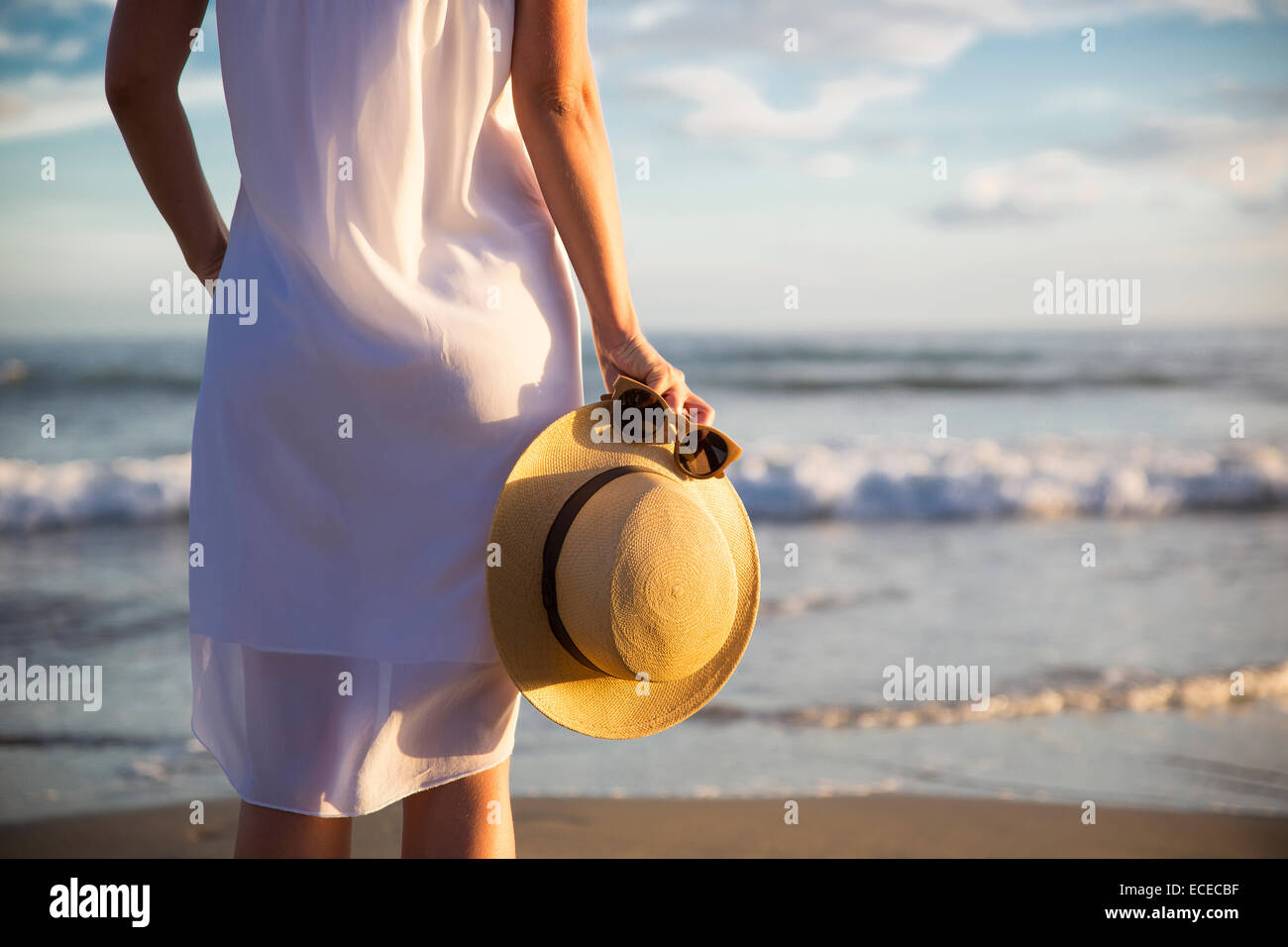 Rear view of woman in white dress standing on beach and holding hat Stock Photo