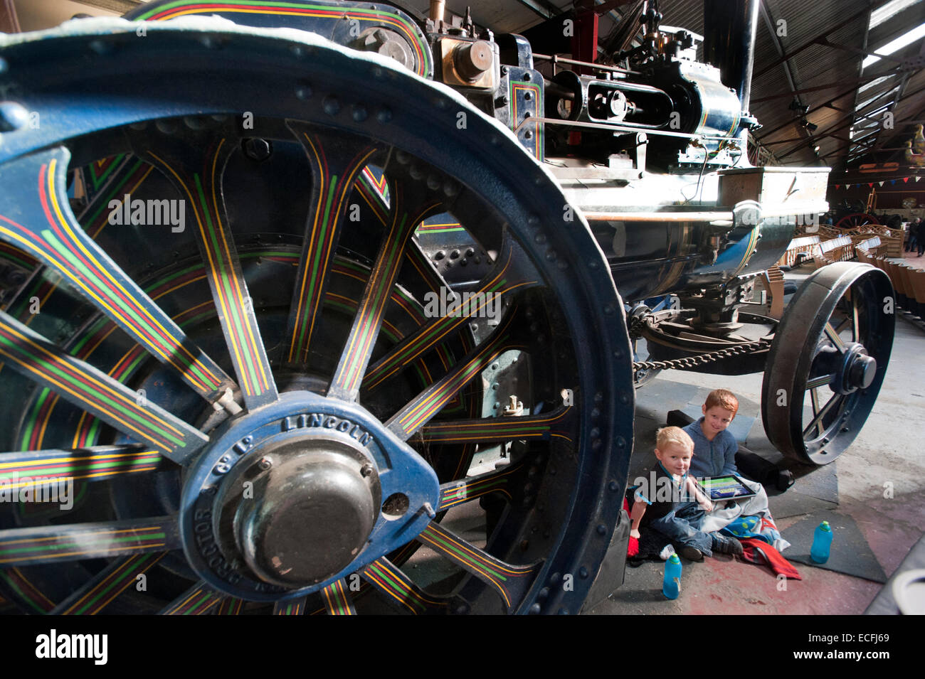 Two young brothers sit under the giant decorative wheels of Traction Engine 'The Champion' built by Ruston Proctor & Co of Lincoln at Strumpshaw Hall Steam Museum near Norwich, Norfolk. The traction engine was on display along with steam rollers, showmans engines and ploughing engines in the exhibition hall. Stock Photo