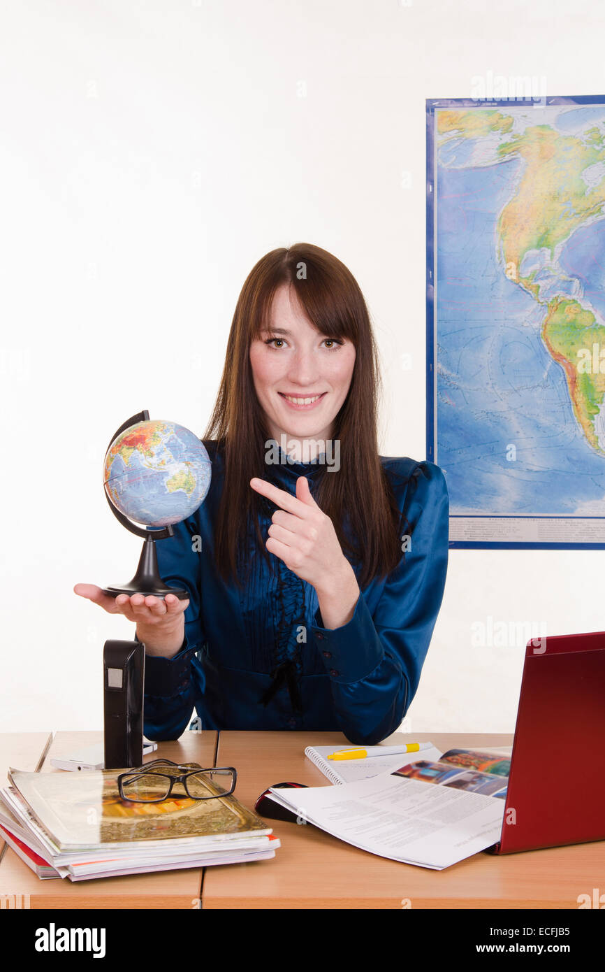 Young geography teacher sitting behind a desk, behind which hangs a map