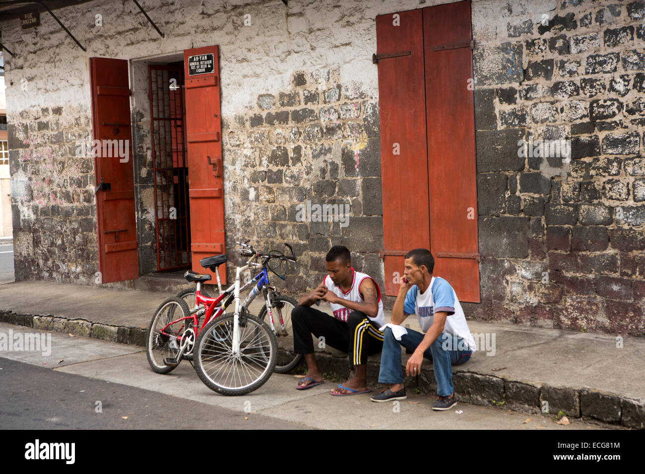 Mauritius, Mahebourg, town centre, men with bikes sat on pavement outside colonial era shop Stock Photo