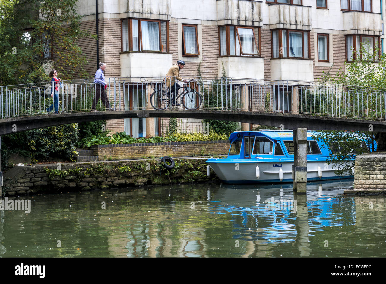 People walk and a man cycles over a footbridge along the River Thames in Oxford, UK. A boat is moored along the river bank. Stock Photo