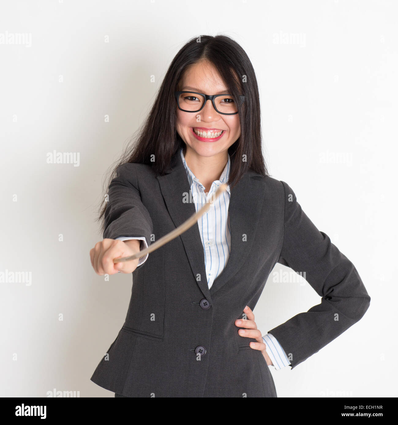 Asian female teacher smiling and holding a stick pointing at camera, standing on plain background. Stock Photo