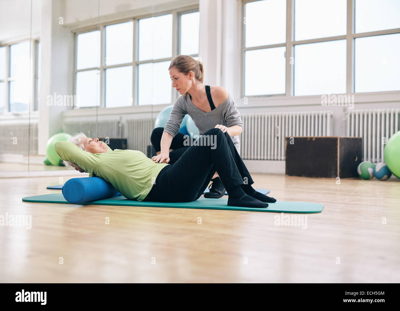Senior woman performing back exercise on a foam roller being assisted by her personal trainer at gym. Physical therapist helping Stock Photo