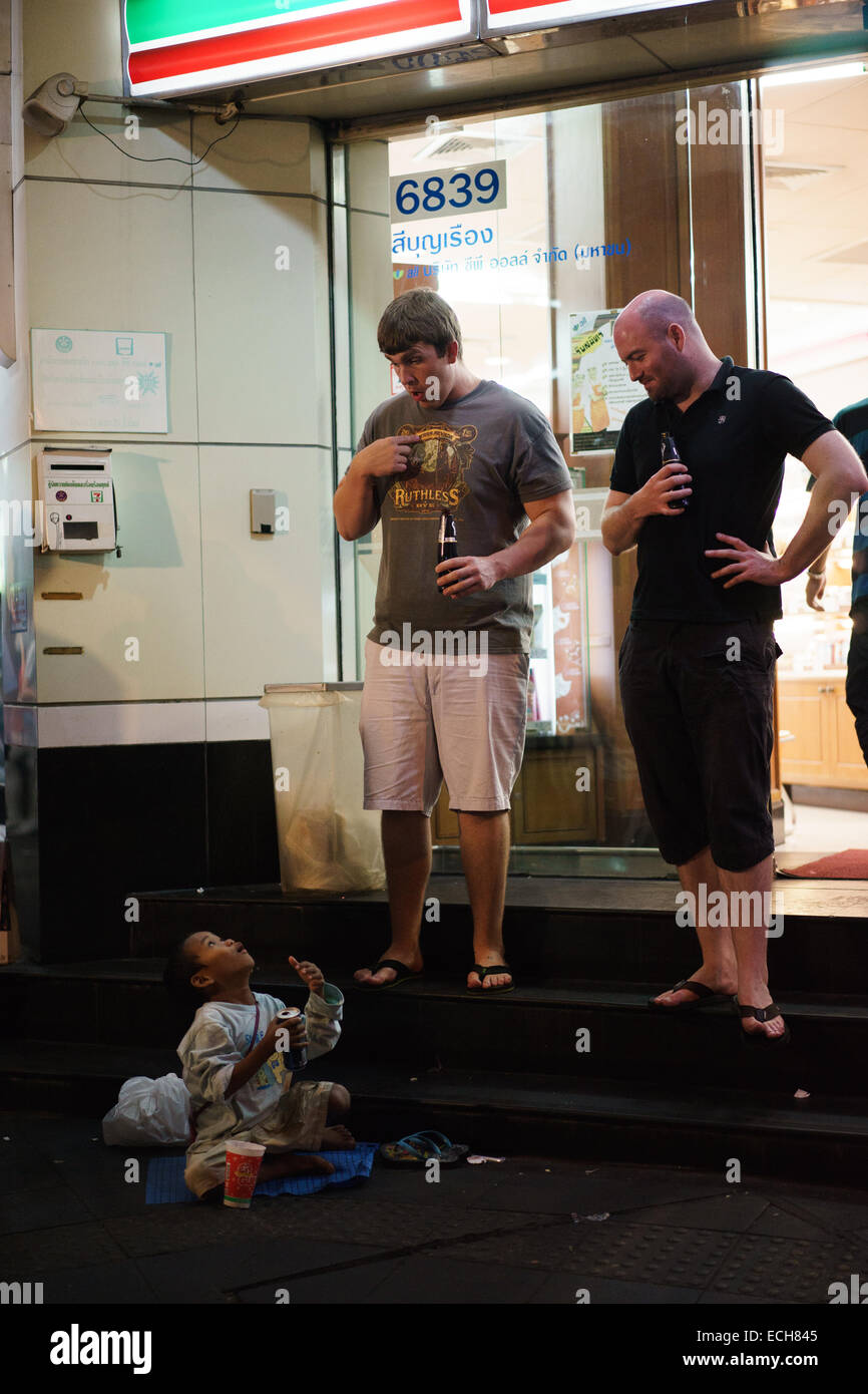 Two western tourists chatting with a child street beggar in front of a seven-eleven shop on Silom road, Bangkok, Thailand. Stock Photo