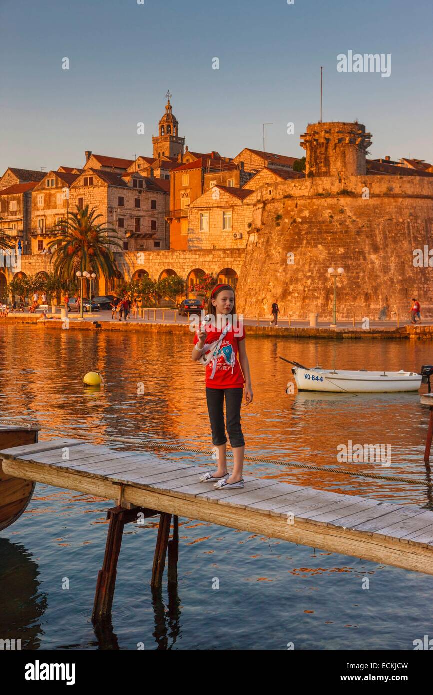 Croatia, Dalmatia, Dalmatian coast, Korcula Island, Korcula, a young girl standing on a wooden bridge in front of a fortified port city at sunset Stock Photo