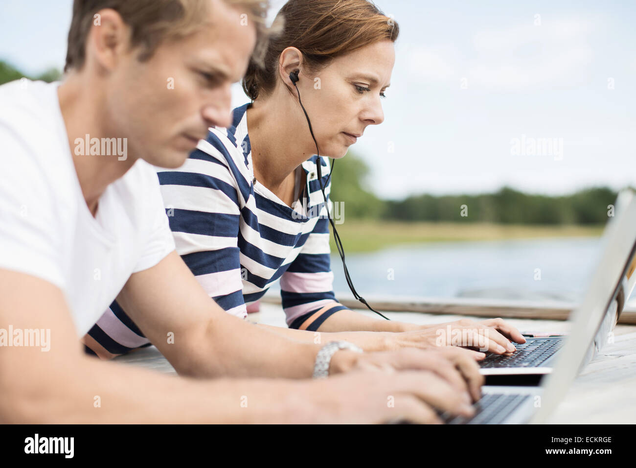Side view of mature couple using laptops while lying on pier Stock Photo