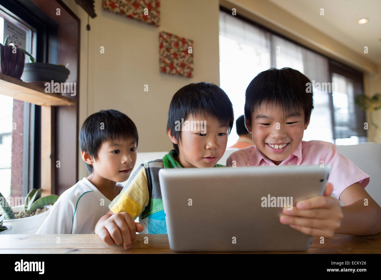 Three boys sitting at a table, looking at a digital tablet, smiling. Stock Photo