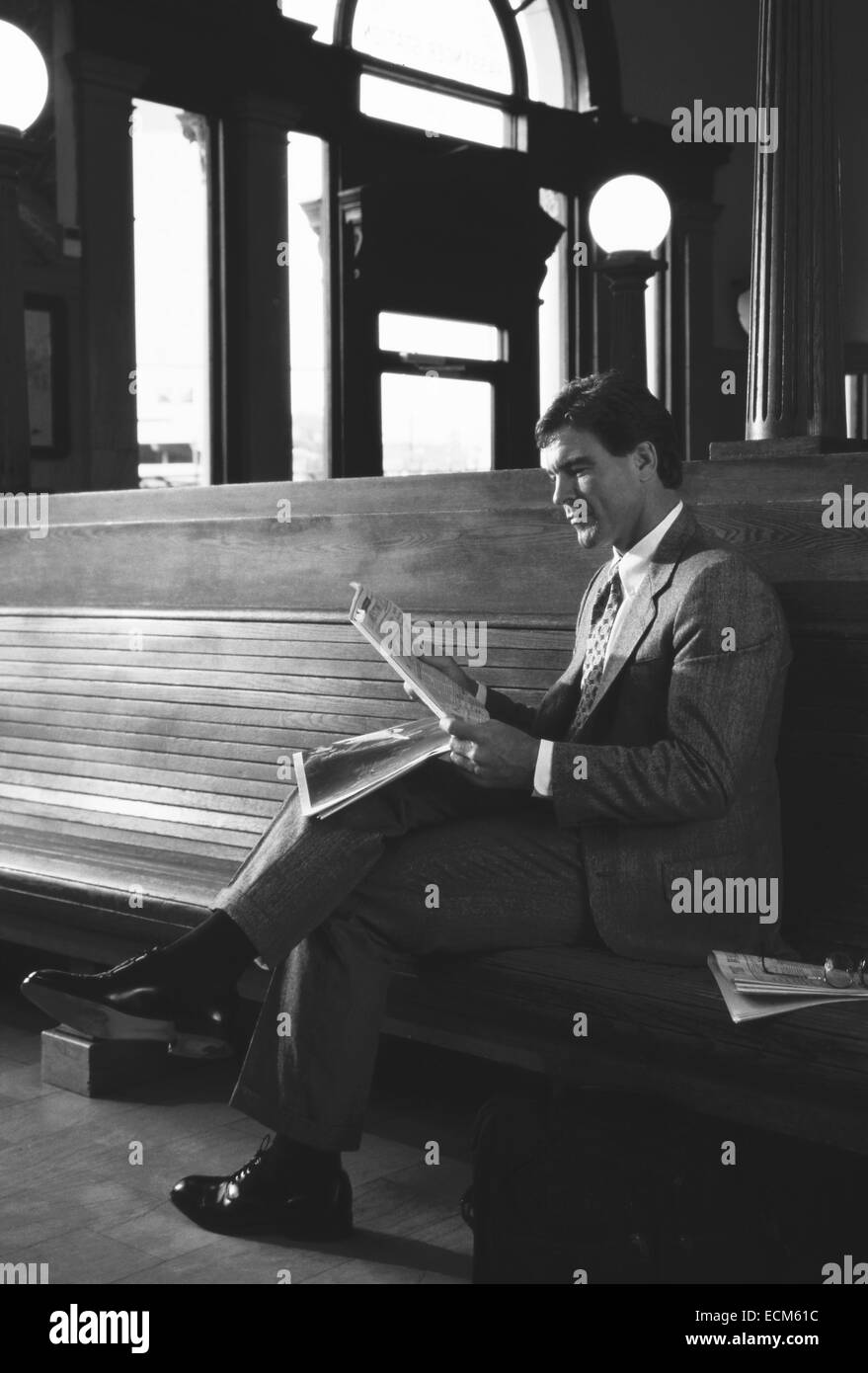 A handsome businessman in a suit reads a newspaper sitting on a wooden bench at a railway train station. Stock Photo