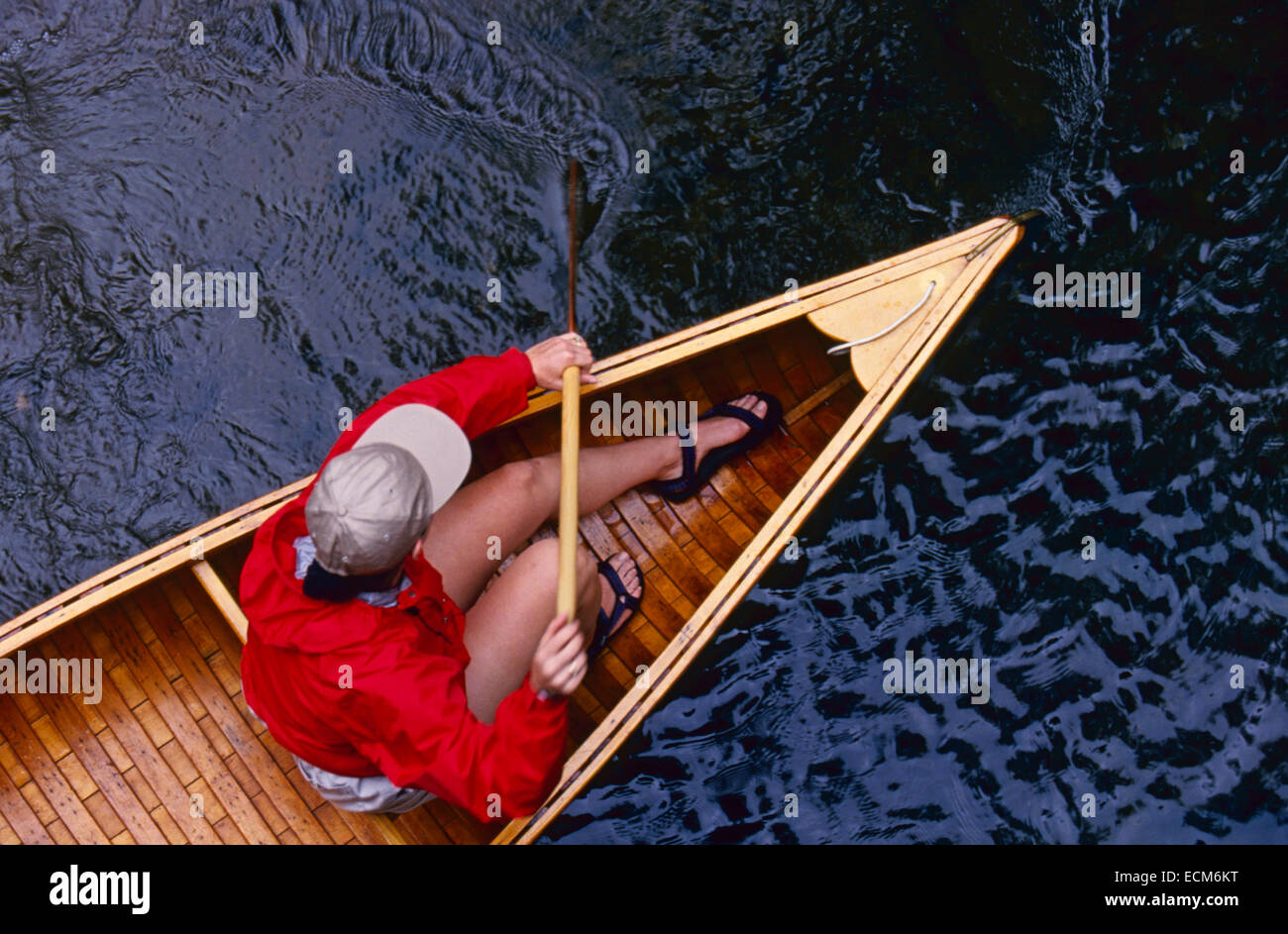 A young woman in a bright red jacket paddles a vintage wooden canoe in a stream Stock Photo