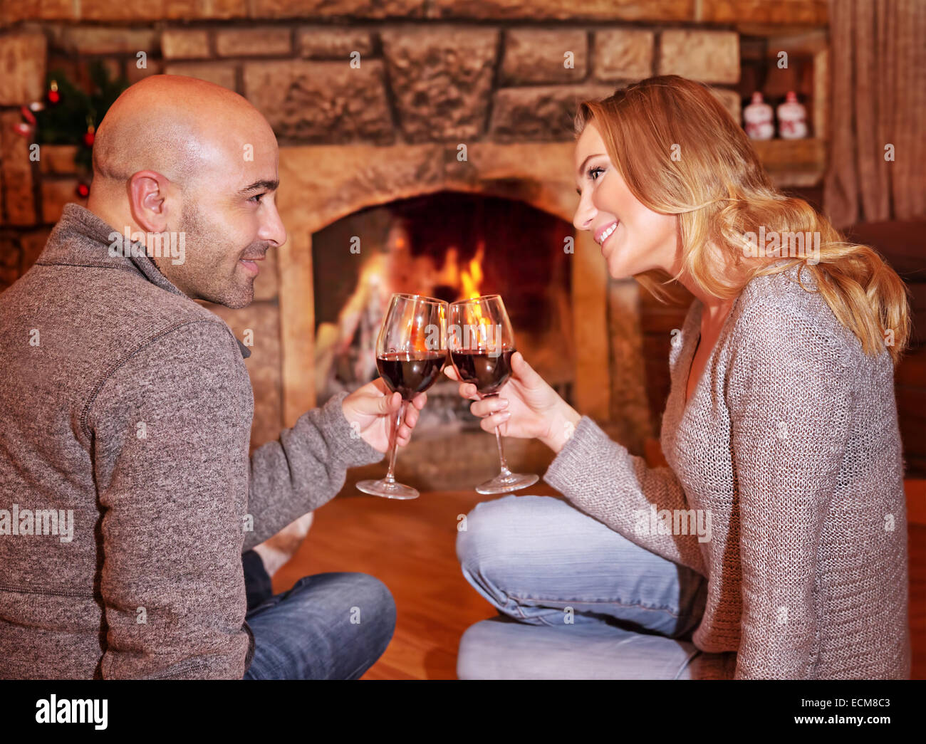 Side view of happy cheerful couple sitting on the floor near fireplace, clinking glasses with tasty red wine Stock Photo