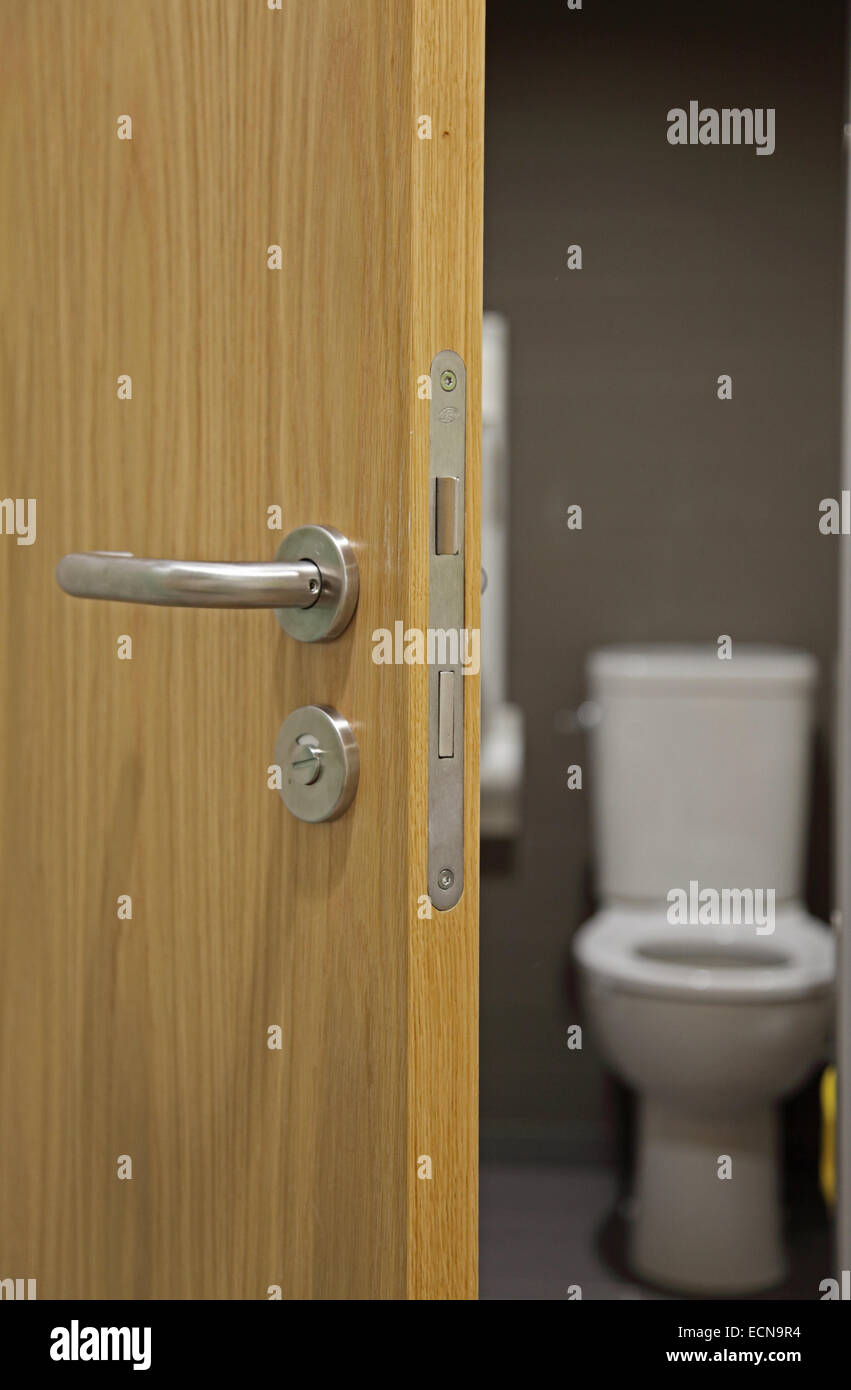 A partially open, modern, oak-finish interior door showing door handle, lock and mechanism showing a toilet beyond Stock Photo