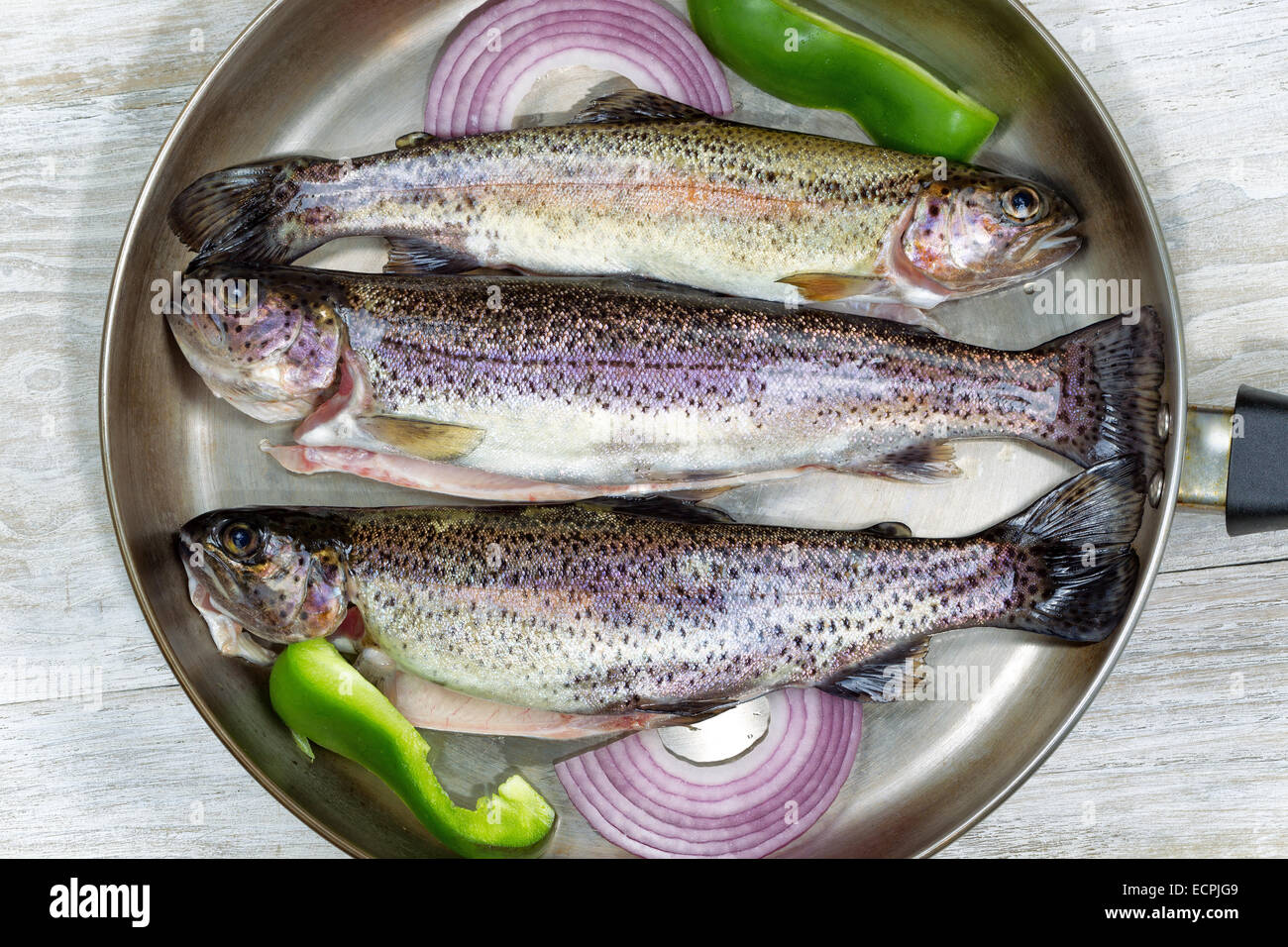 Top view of fresh wild trout in frying pan with onion and green peppers on rustic white wood. Stock Photo