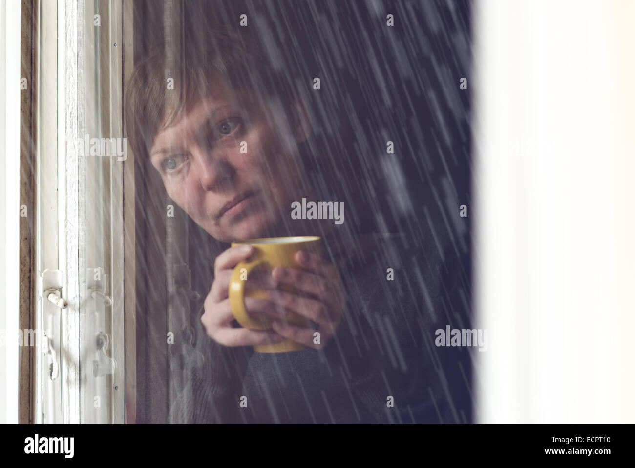 Lonesome woman drinking cup of coffee by the window of her living room, looking out at rain falling with a sad look on her face Stock Photo