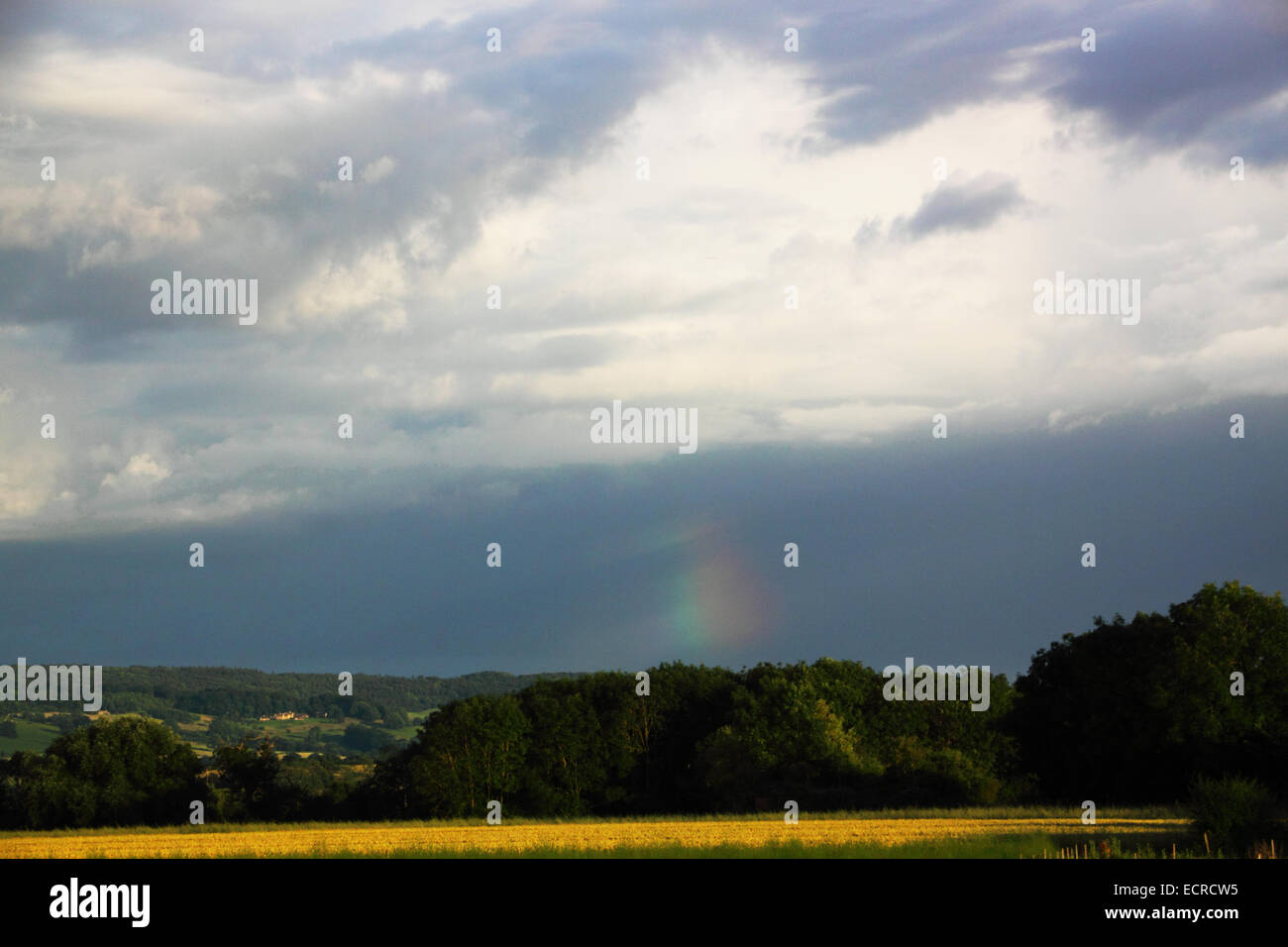 A part of a rainbow above fields on the horizon with dark clouds above. Stock Photo