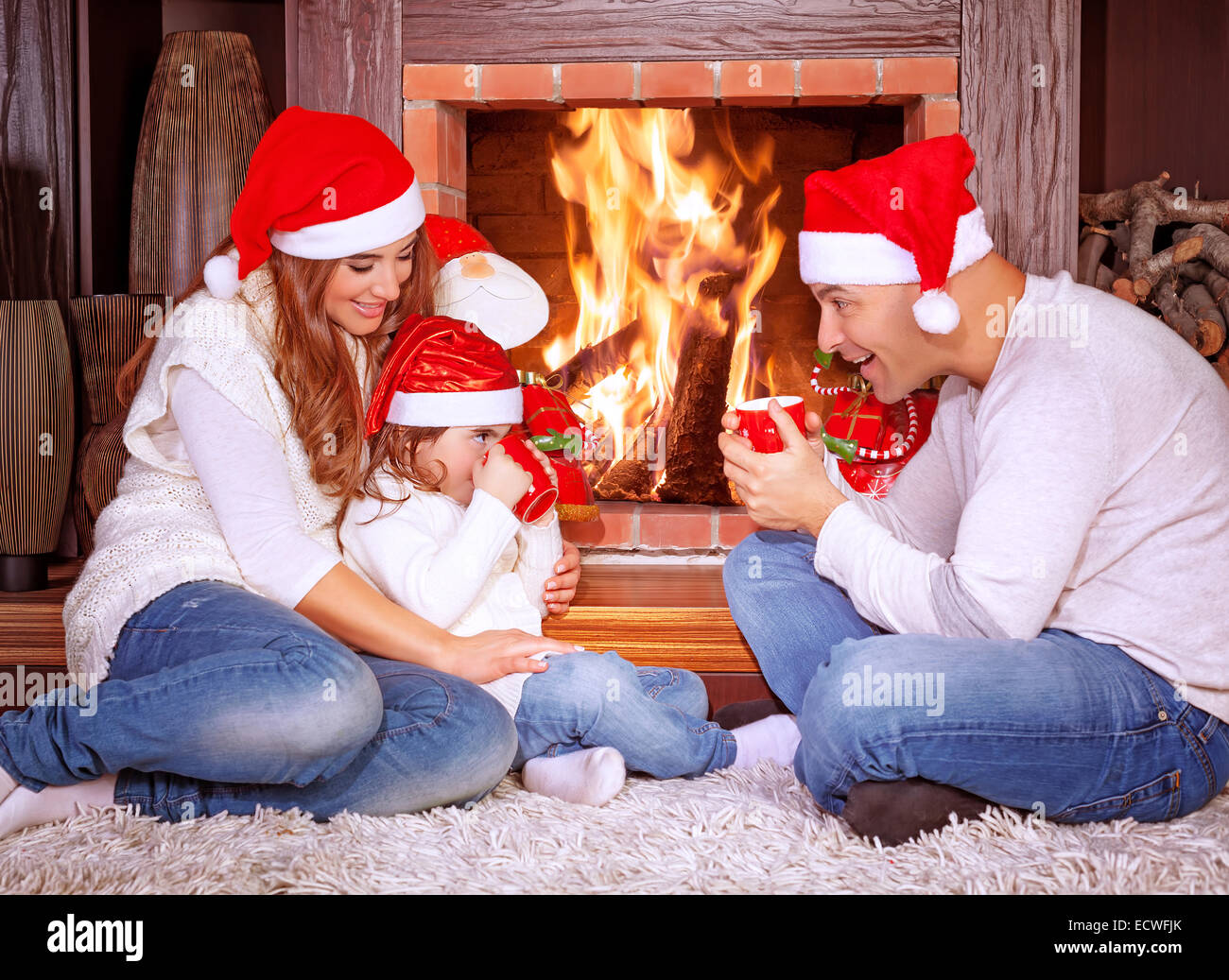 Happy family by fireplace, parents with baby girl wearing Santa Claus hats, sitting relaxed in ski resort chalet Stock Photo