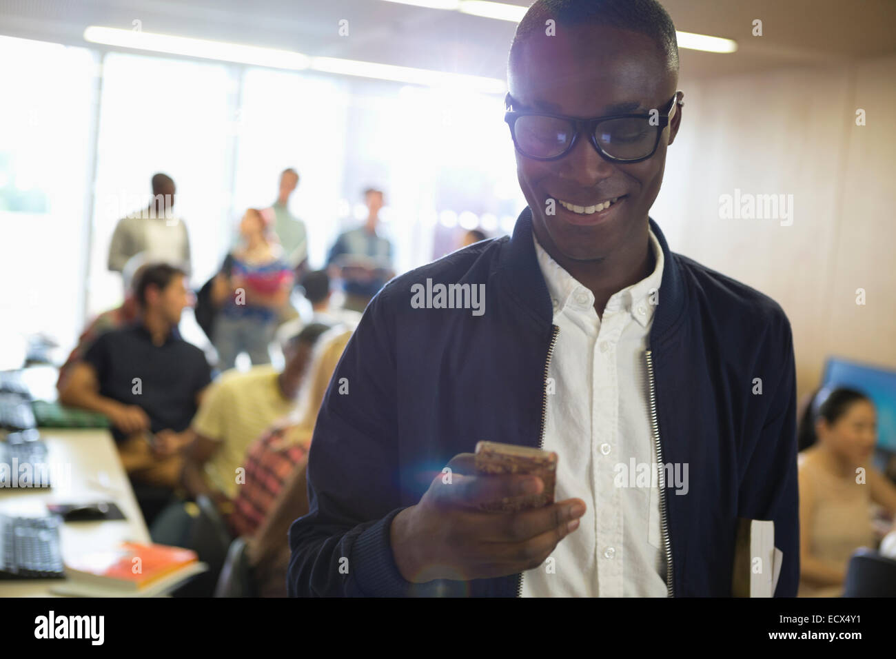 Smiling male student in eyeglasses looking at smart phone, with other students in background Stock Photo