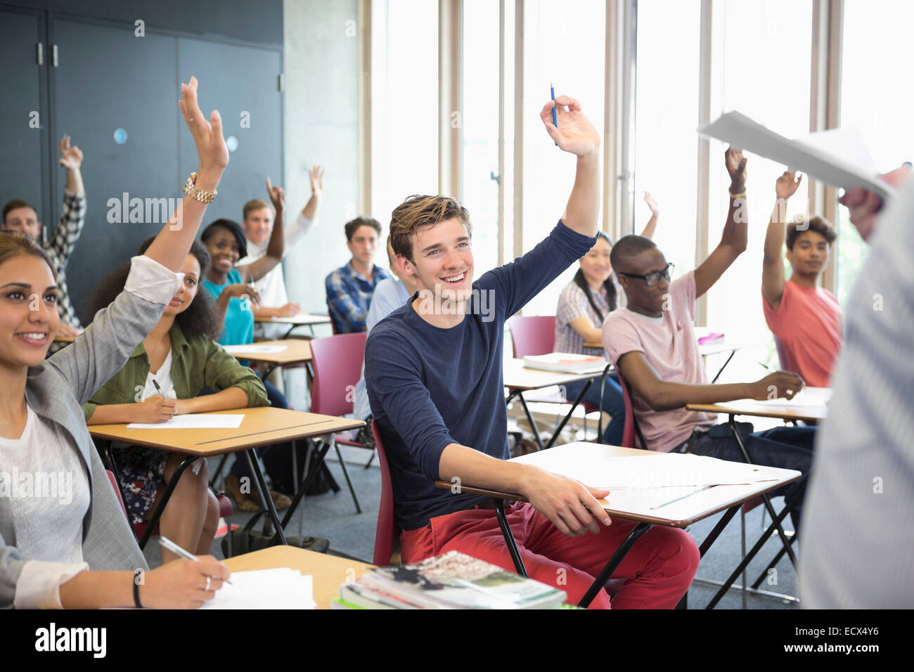 View of smiling students sitting at desks in classroom with arms raised Stock Photo