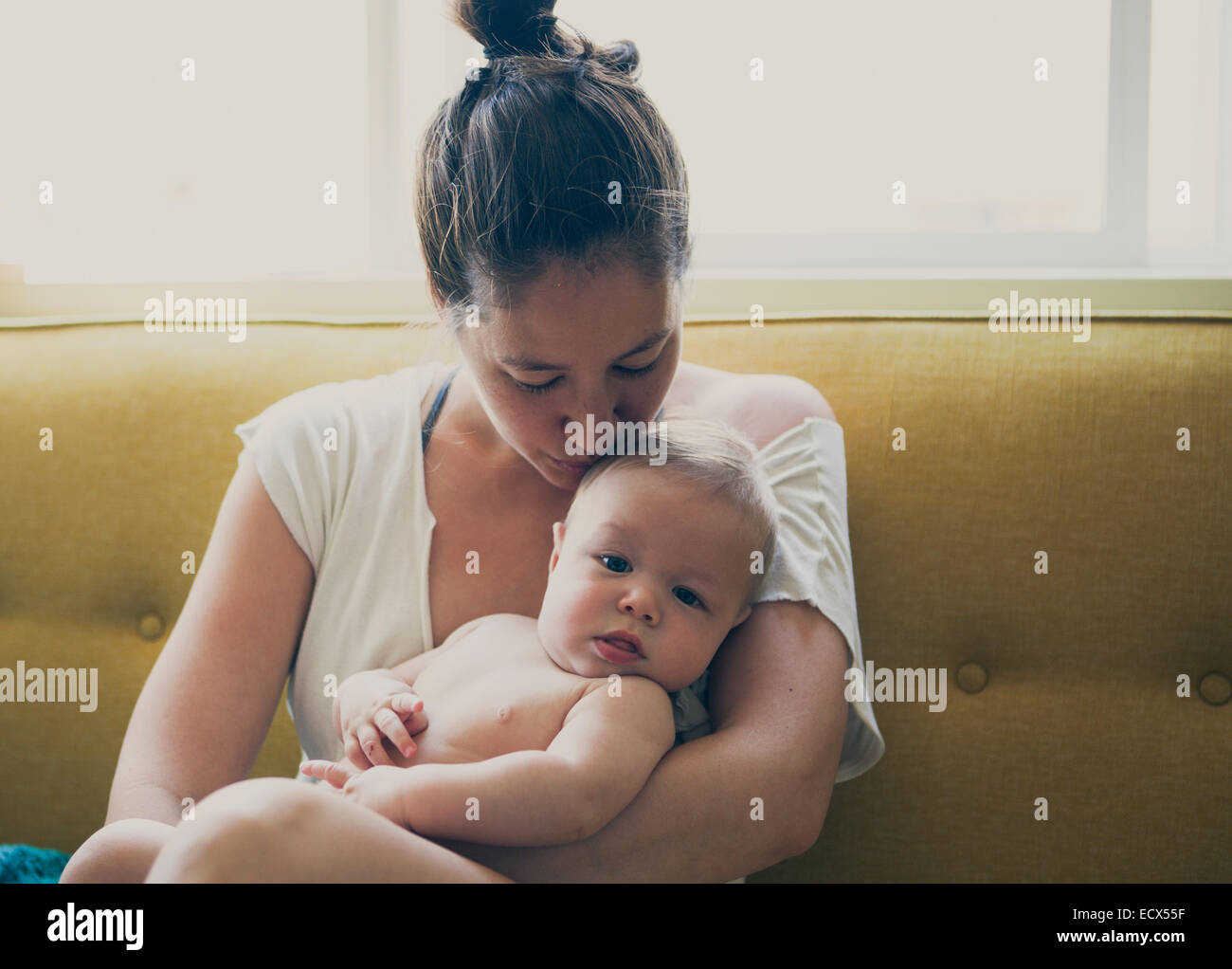 Portrait of mother holding and kissing baby, sitting on sofa by window Stock Photo