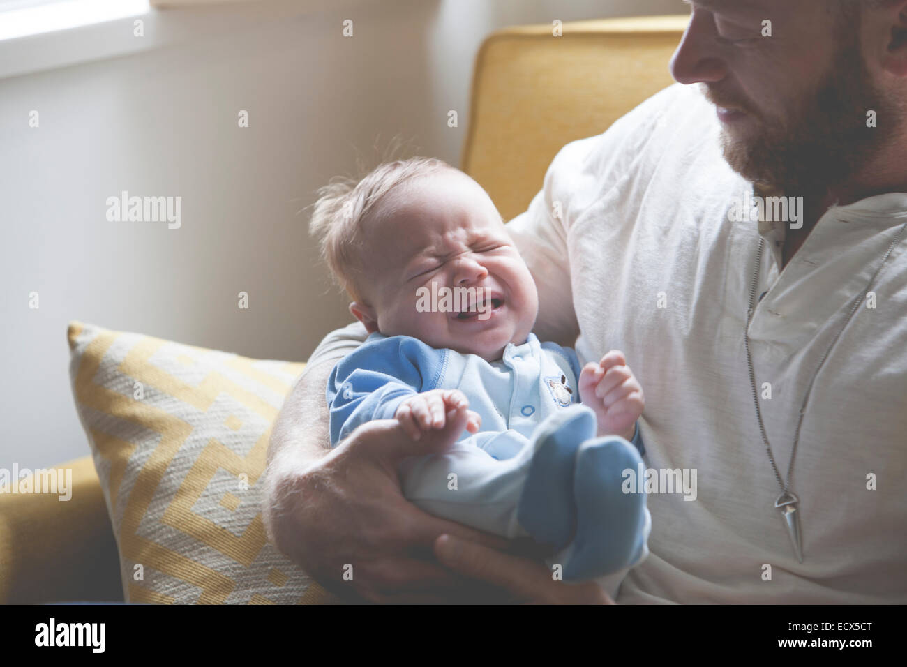 Father holding and looking at crying baby, sitting on sofa Stock Photo