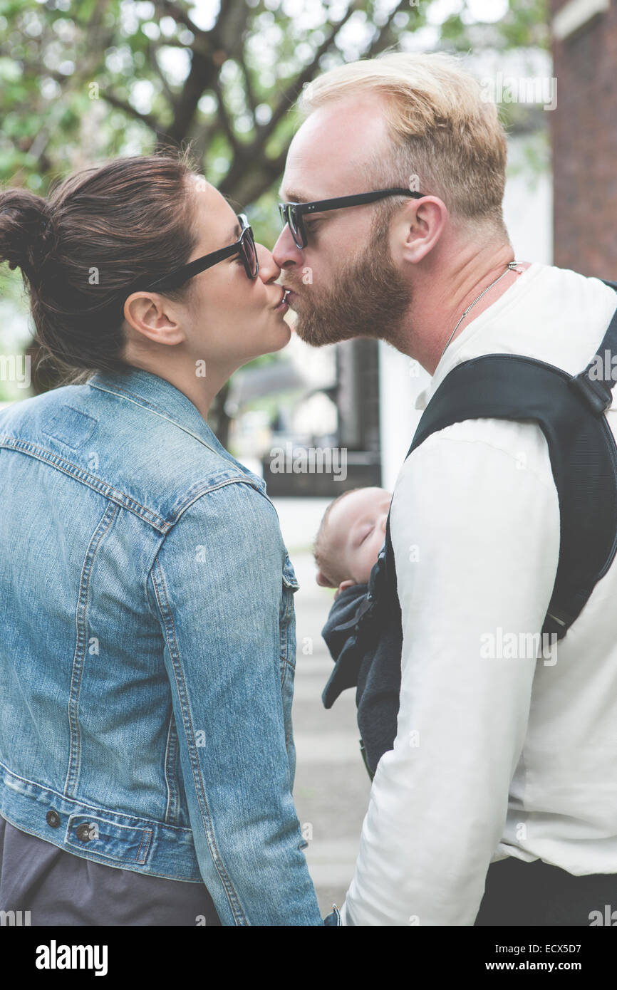 View of couple in sunglasses kissing and holding baby in city streets Stock Photo