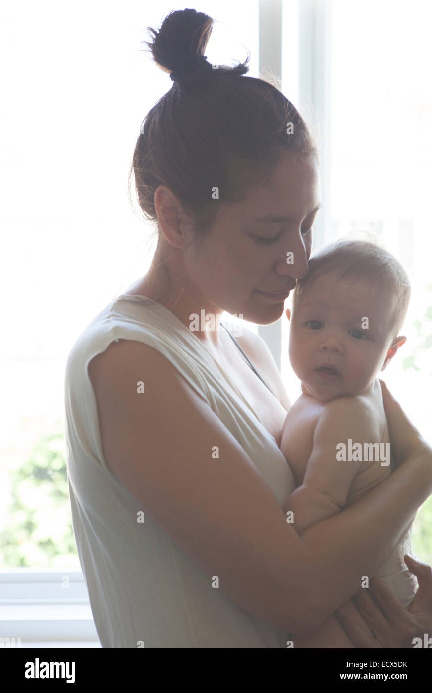 Portrait of mother holding baby, standing beside window with eyes closed Stock Photo