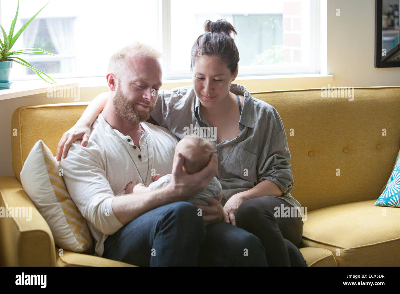 Parents smiling and holding little baby, sitting on yellow sofa Stock Photo