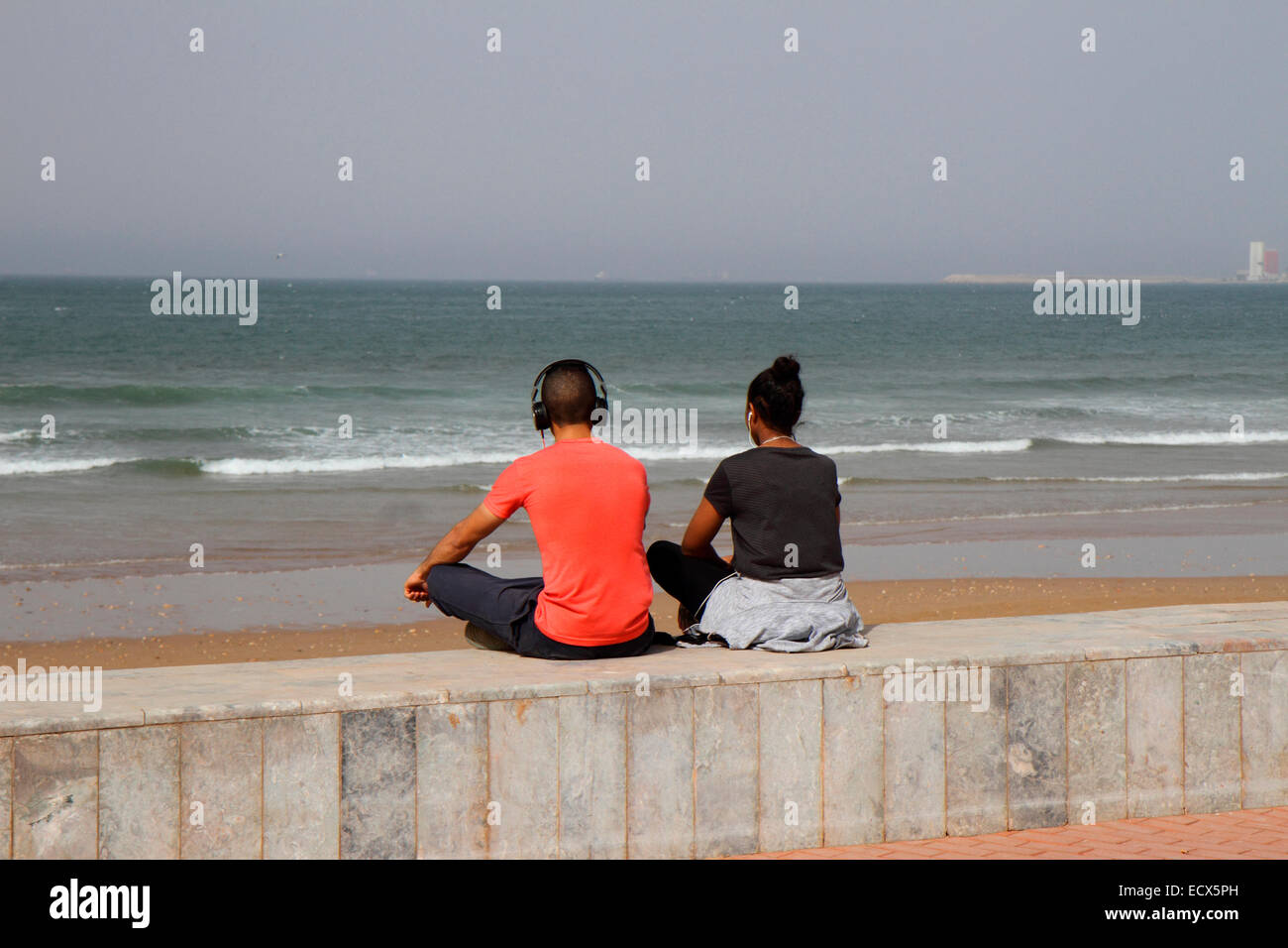 Young man and woman sitting on wall looking out to sea and wearing head phones Stock Photo